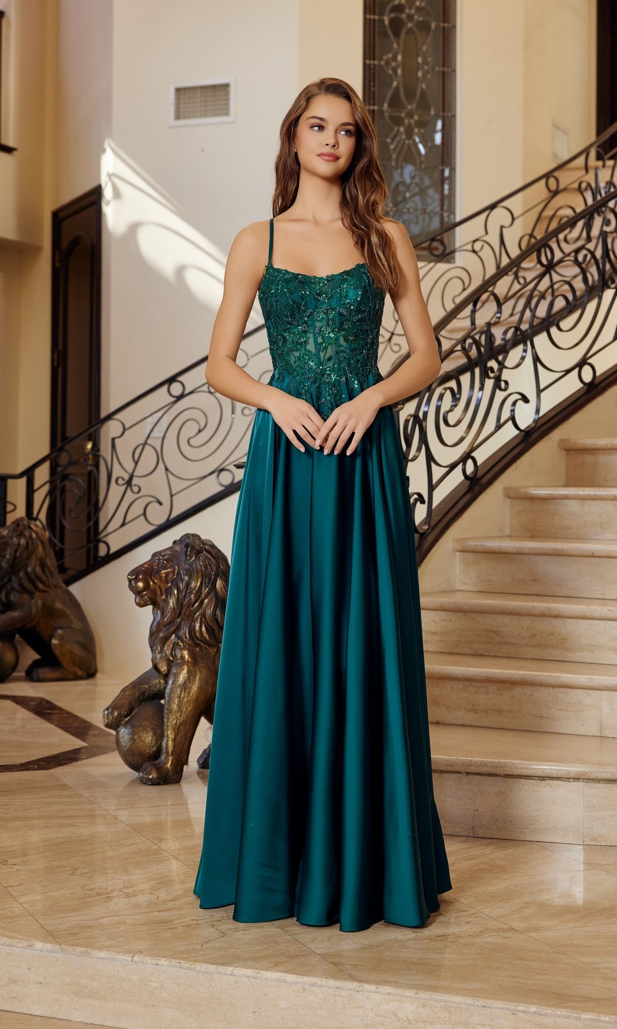 An elegant woman in the Nox Anabel A1725 long prom dress in emerald, featuring a satin A-line skirt, stands on a marble floor before a staircase with ornate railings and two lion statues.